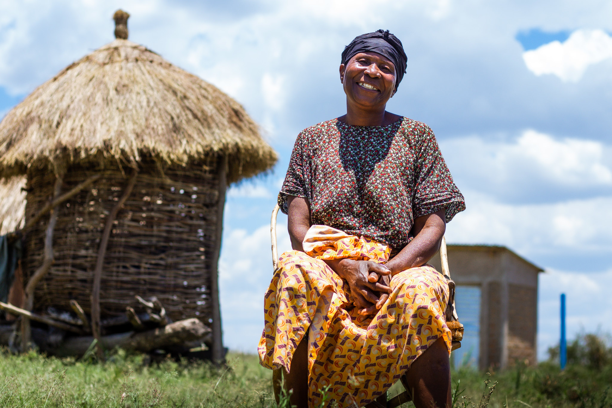 Tanzanian woman in colorful dress smiles sitting in front of a backdrop of her newly built latrine and blue skies.